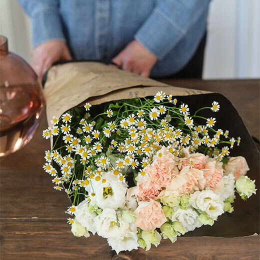 En flott bukett med tre ulike blomster i hvitt og ferskentoner innpakket i papir